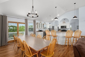 A spacious dining area with a long table and wooden chairs, a kitchen with white cabinets and modern appliances, and a balcony with a view of greenery.