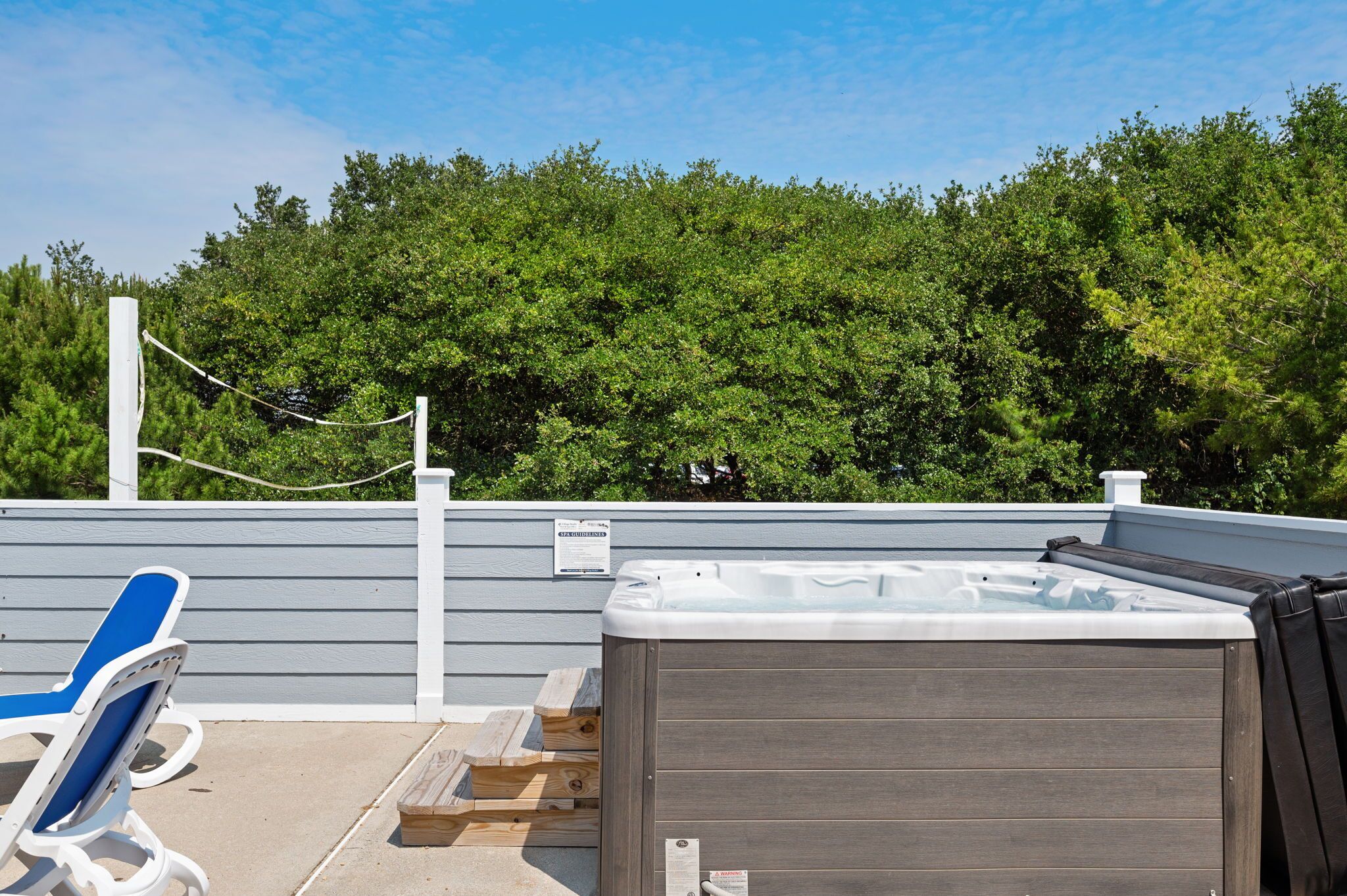 A hot tub with a wooden deck and lounge chairs, surrounded by a wooden fence and trees.