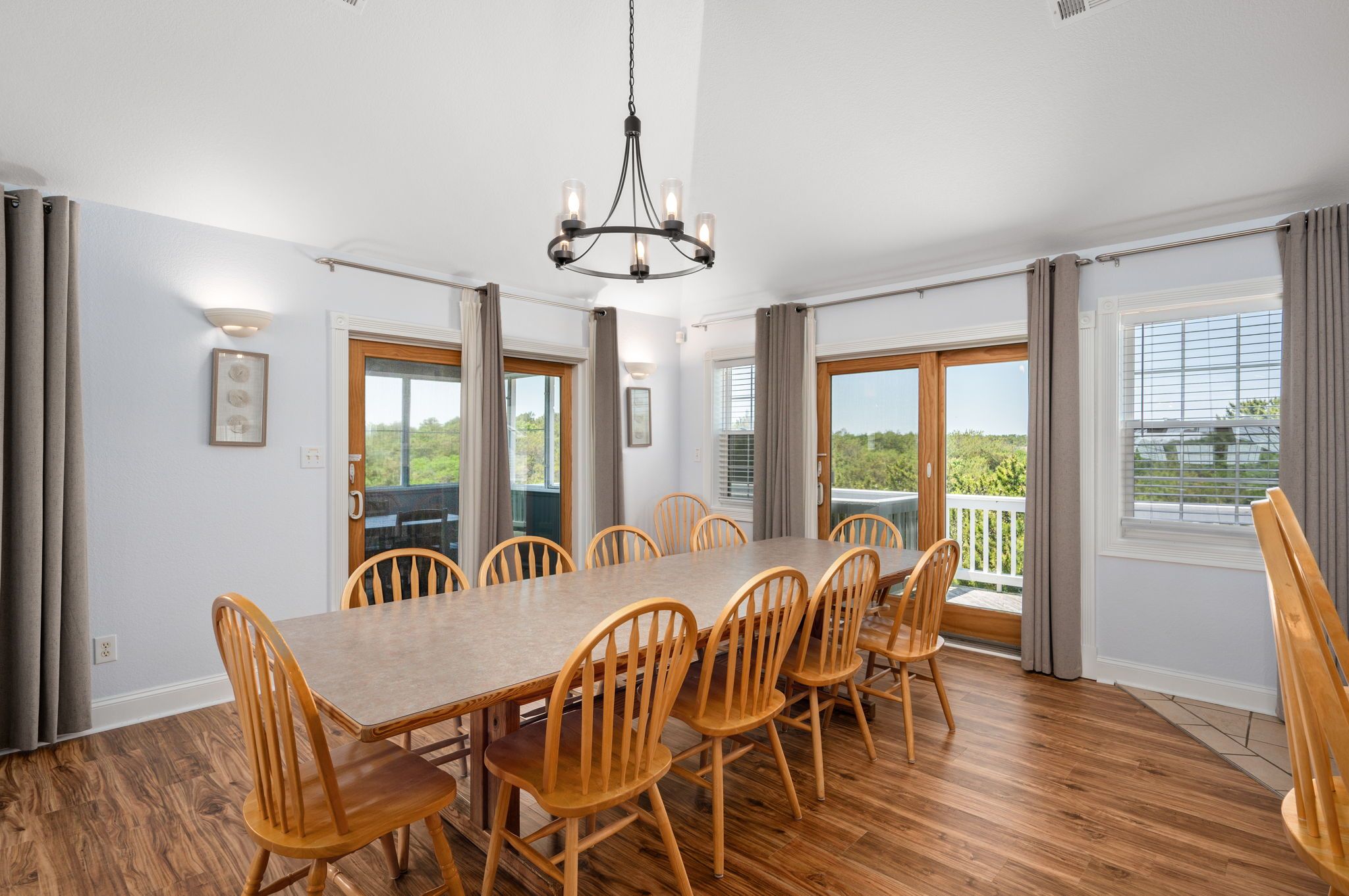A dining room with a long wooden table and chairs, a chandelier, and large windows with curtains.