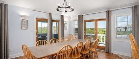A dining room with a long wooden table and chairs, a chandelier, and large windows with curtains.