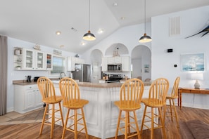 A spacious kitchen with white cabinetry, a central island with bar stools, and a dining area with a wooden table and chairs. - Heaven Sent: Oceanside, private pool, hot tub, rec room and EASY beach access! (Corolla)