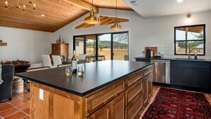 Une cuisine moderne dotée d’un grand îlot central, de meubles en bois et d’un tapis rouge. On y trouve un coin repas avec une table et des chaises, et une vue sur l’extérieur grâce à de grandes fenêtres.