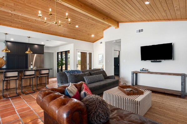 A spacious living area with a leather sofa, a television mounted on the wall, and a kitchen area with dark cabinets and a stainless steel refrigerator.