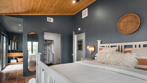Une chambre moderne avec un grand lit, un plafond en bois, un luminaire fixé au mur et une salle de bain visible à travers une porte ouverte.