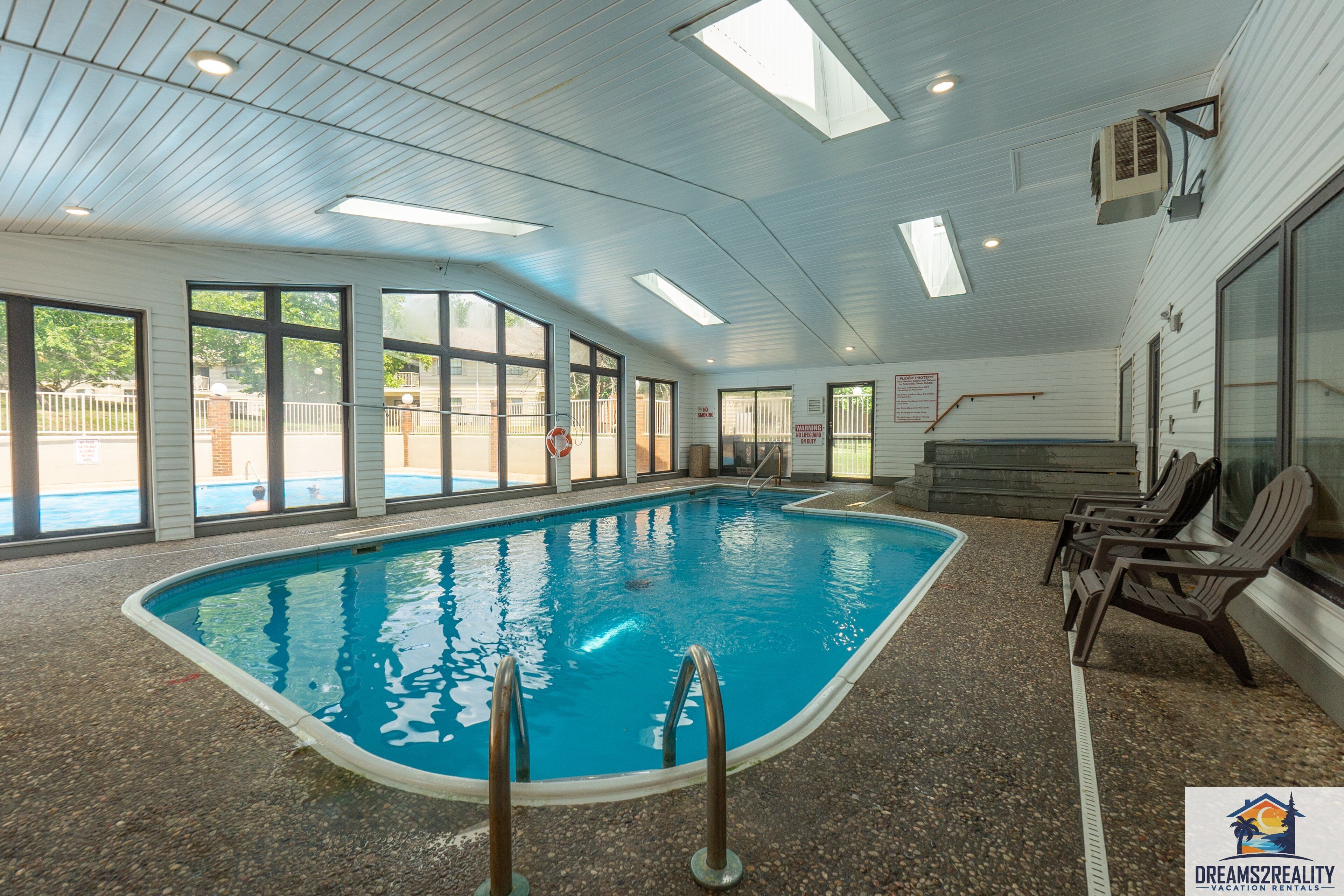 Una piscina interior con paredes de vidrio transparente, piso de baldosas y algunas sillas de descanso.
