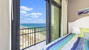A bedroom with a large sliding glass door leading to a balcony with a view of the beach and ocean.
