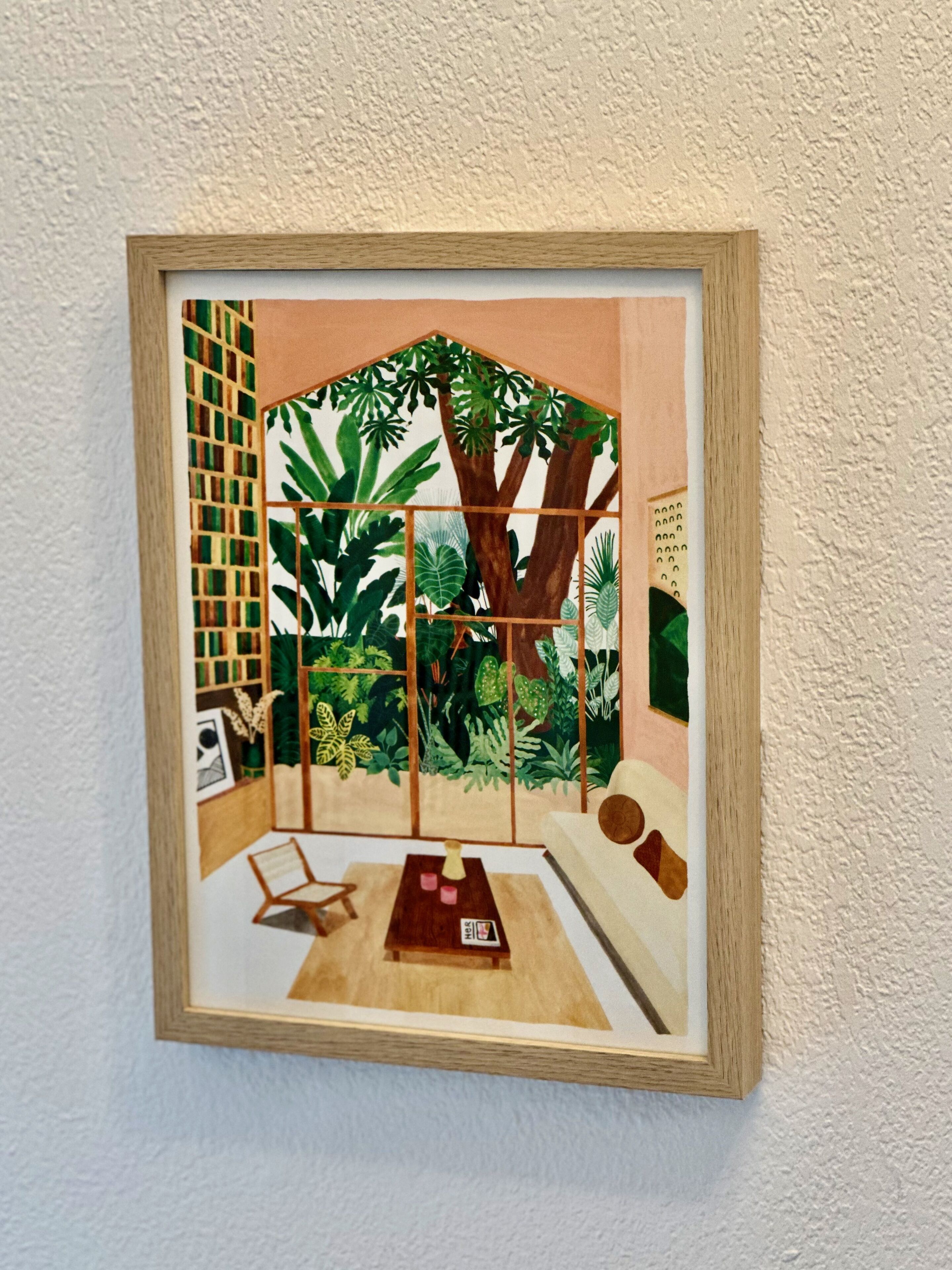 A framed artwork depicting a room with a large tree, a sofa, a small table, and a chair.