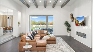 A spacious living room with a sectional sofa, a flat-screen TV, and a large window overlooking a pool and outdoor seating area. - Gated Estate Near Troon N. Pool, Hot-tub, Pickleball, Frisbee Golf, Bar & Arcade (Scottsdale)