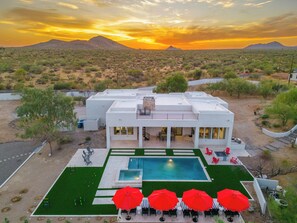 Un edificio moderno con piscina y asientos exteriores, rodeado de vegetación desértica y montañas al fondo.