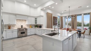 A modern kitchen with white cabinetry, a central island with a sink, and a large window providing a view of the outdoors. - Gated Estate Near Troon N. Pool, Hot-tub, Pickleball, Frisbee Golf, Bar & Arcade (Scottsdale)