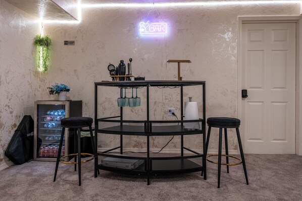 Une pièce avec une table ronde en métal noir, deux tabourets noirs, un panneau lumineux néon indiquant « SYBAR », une petite plante et un réfrigérateur rempli de boissons.