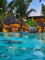 Una piscina con agua cristalina, rodeada de palmeras y construcciones con techo de paja.