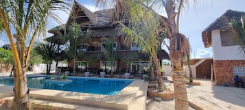 Um resort com piscina, palmeiras e um edifício com telhado de palha.