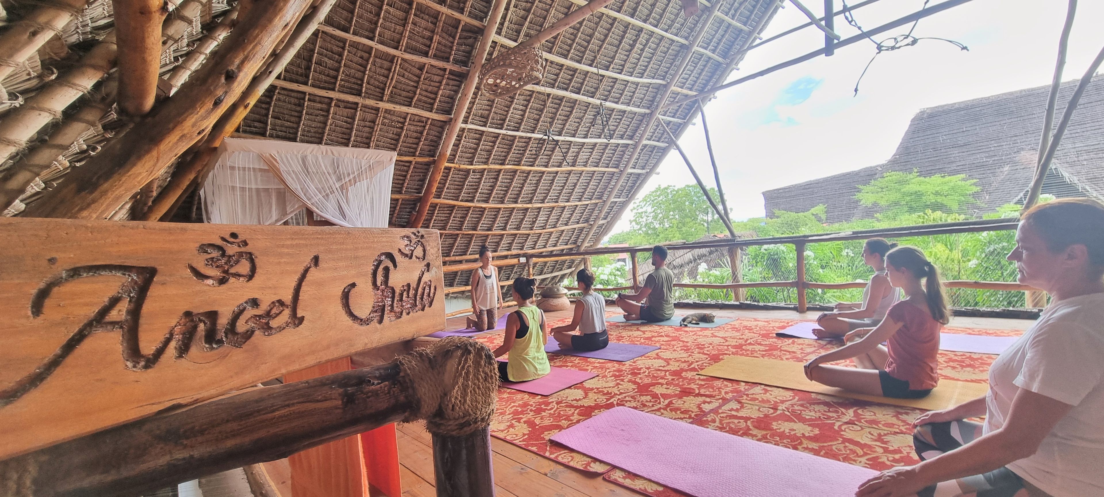 A wooden sign with 'Angel' inscribed, a thatched roof structure, and people practicing yoga on mats.
