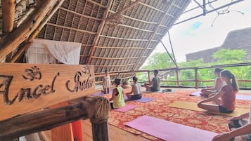 Een houten bord met de tekst 'Angel', een gebouw met rieten dak en mensen die yoga doen op matten.