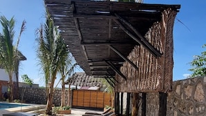A sunlit outdoor seating area with wooden loungers under a thatched roof, surrounded by palm trees and a stone wall. - Salama & Mbuyuni Bungalows (Matemwe)