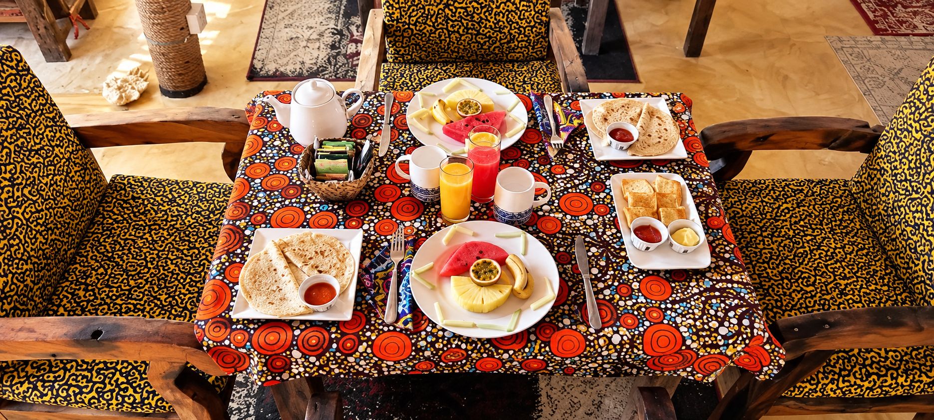 Une table dressée avec une variété d’aliments pour le petit-déjeuner, notamment des galettes, des fruits et des boissons, et recouverte d’une nappe à motifs.