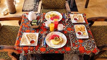 Une table dressée avec une variété d’aliments pour le petit-déjeuner, notamment des galettes, des fruits et des boissons, et recouverte d’une nappe à motifs.