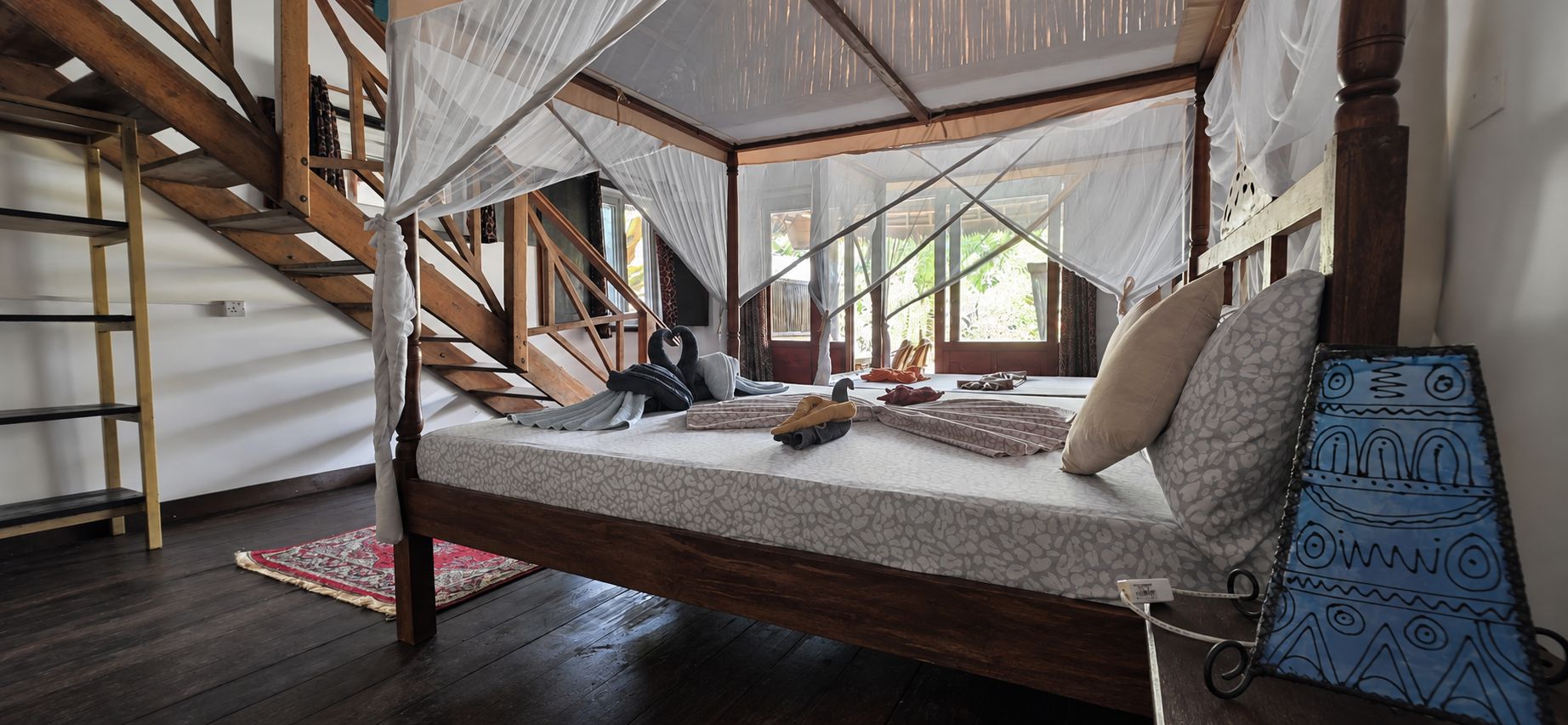 Una cama con dosel, vigas de madera, una escalera, un tapete rojo y un colgador azul con estampado.