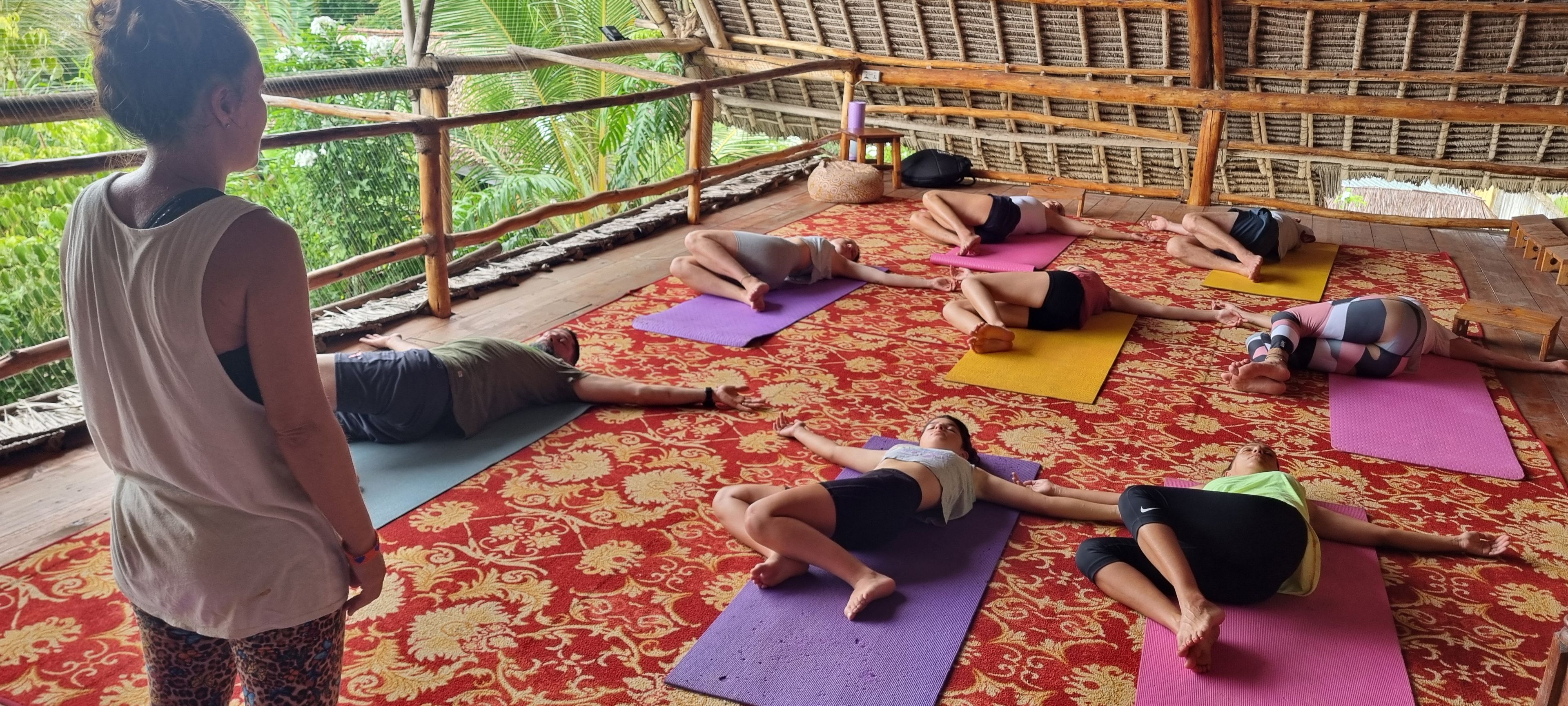 Un grupo de personas practicando yoga sobre colchonetas en una habitación con alfombra estampada y techo de paja.