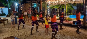 Un grupo de personas bailando en una playa arenosa de noche, con una estructura de techo de paja y luces colgantes al fondo.