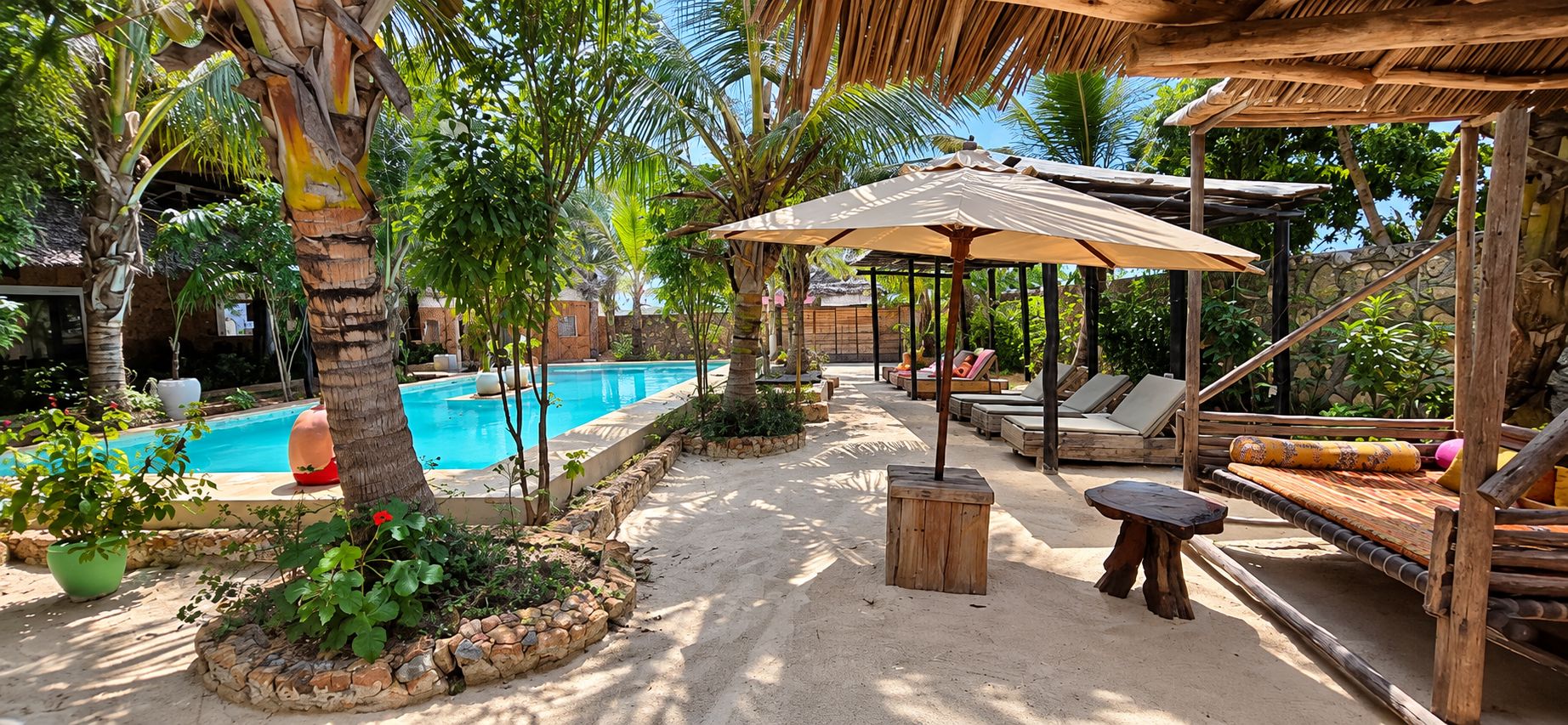 Zona junto a la piscina con sillones de descanso y sombrillas, rodeada de vegetación tropical y un muro de piedra.