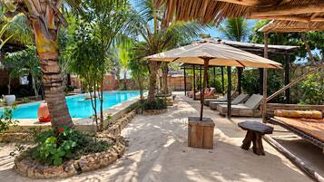Zona junto a la piscina con sillones de descanso y sombrillas, rodeada de vegetación tropical y un muro de piedra.