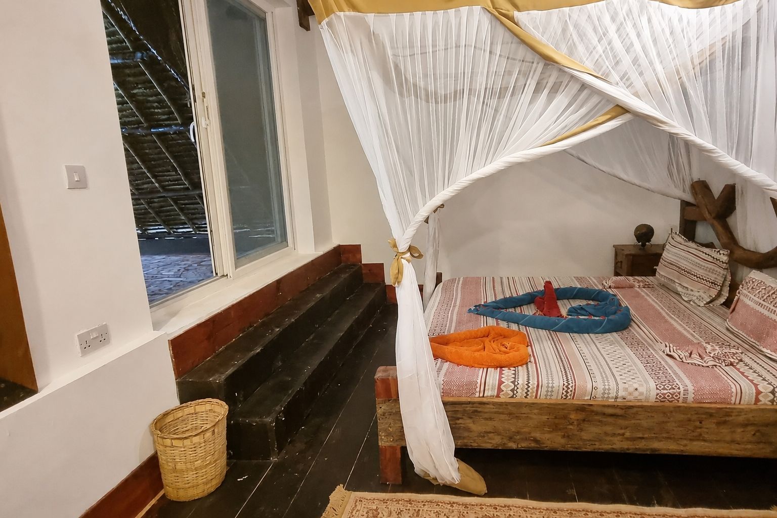 A room with a bed, a window, a wooden staircase, and a woven basket.