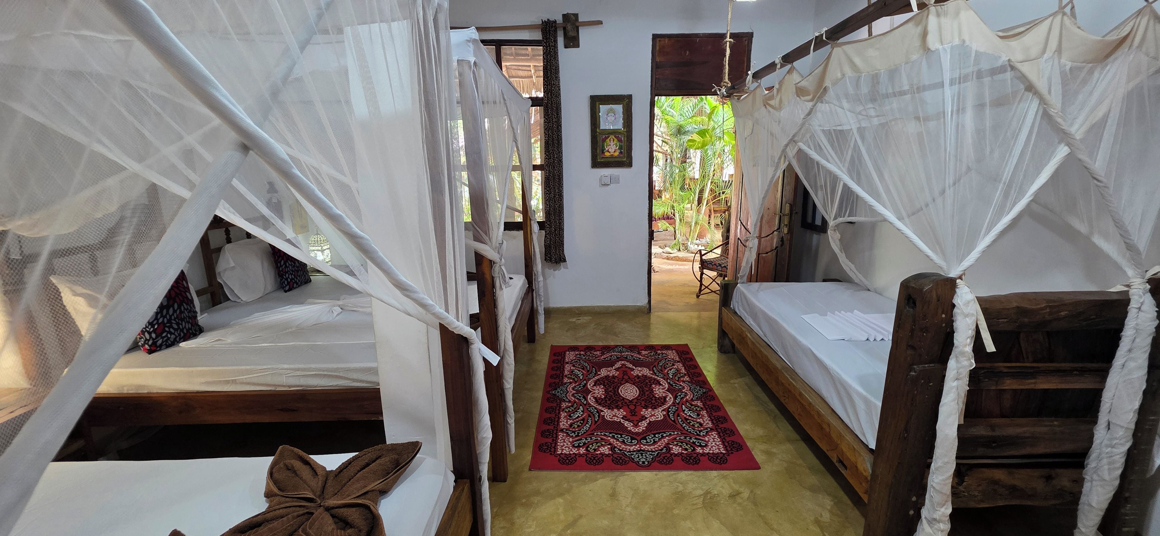A corridor with two wooden beds, each with a canopy, leading to a door. A red patterned rug is on the floor.
