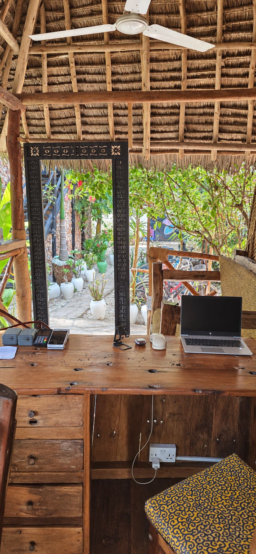 A wooden desk with a laptop, a mobile phone, and a cash register. There is a mirror with a decorative frame, and a patterned cushion on the floor.