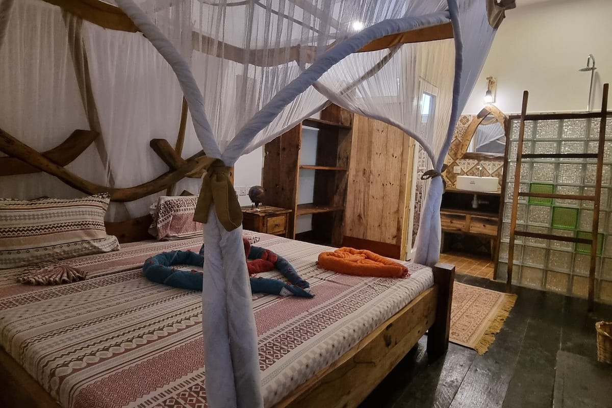 Habitació amb un llit, un panell de capçalera de fusta, una mosquitera i vista al bany amb dutxa.
