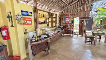 Un interior rústico con mostrador de madera, estanterías con diversos objetos, un extintor y un tejado de paja.