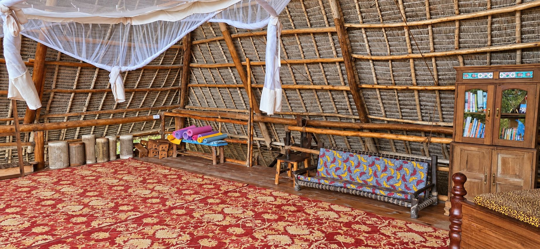 A room with a thatched roof, a red patterned carpet, a wooden cabinet with books, and a bench with a colorful cushion.