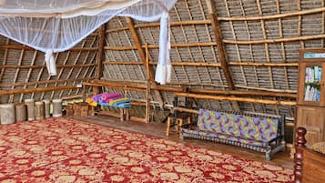Una habitación con techo de paja, alfombra roja con estampado, un armario de madera con libros y un banco con cojín colorido.