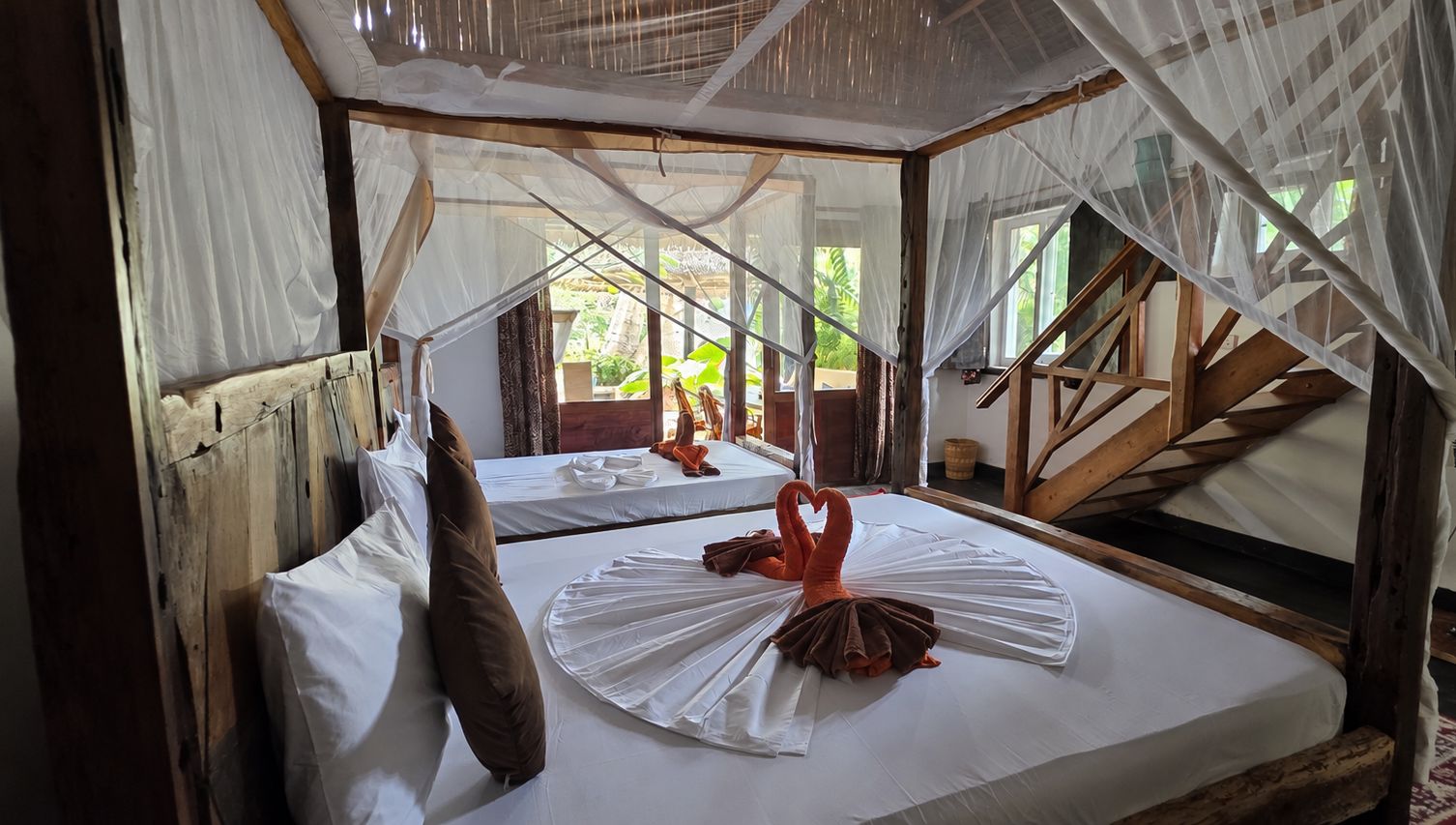 A room with two beds, each with white linens and a brown pillow. There is a wooden staircase and a window with a view of greenery outside.