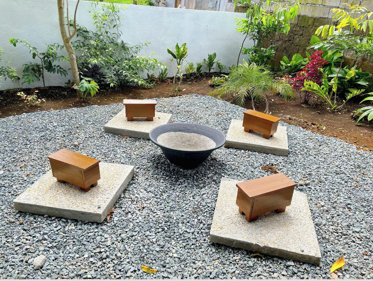 A garden area with a central fire pit surrounded by four square stone platforms, each with a wooden stool.