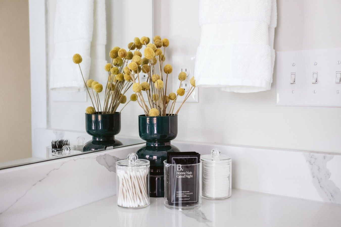 A bathroom vanity with a mirror, two dark green vases with dried yellow flowers, a container of cotton swabs, and a bottle labeled 'Bonne Nuit Good Night.'