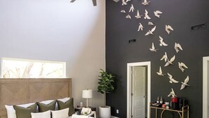 Un dormitorio moderno con una cama grande, un ventilador de techo, un adorno de pájaro en la pared y una silla de vidrio.