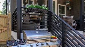 Una parrilla al aire libre con utensilios, una silla de mimbre y macetas colgantes.