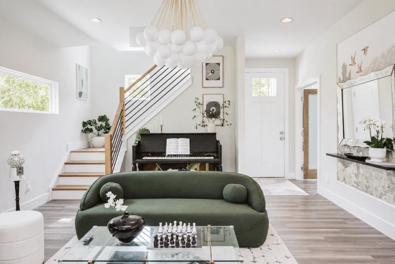 A modern living room with a green sofa, a piano, a staircase, and a chandelier.
