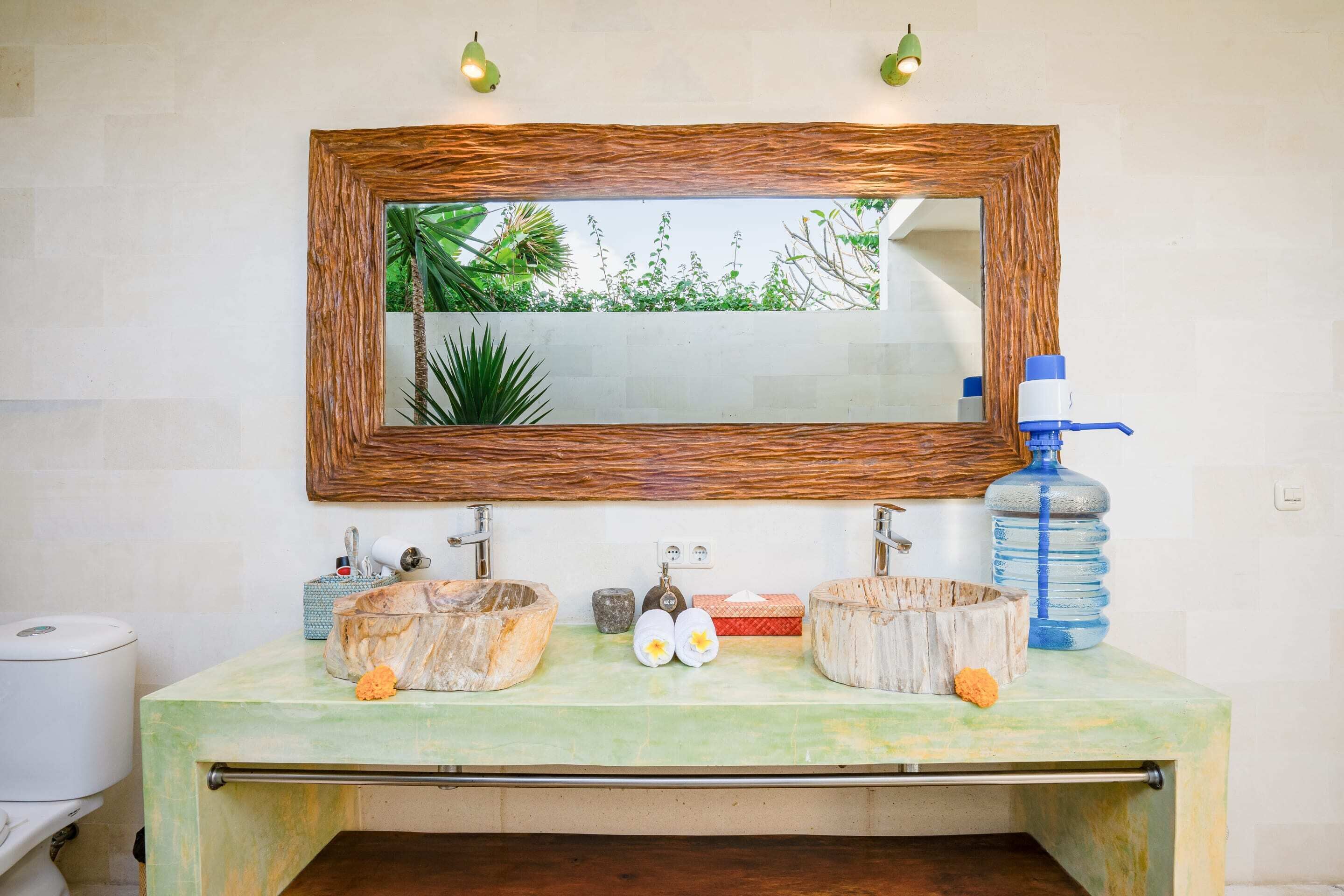 A bathroom with a rustic wooden-framed mirror, two sinks with natural stone bowls, a water dispenser, and a small plant.