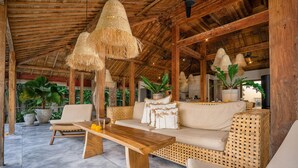 Un espacio exterior techado con mesa de madera y sofá tejido, rodeado de plantas en macetas y techo de paja.