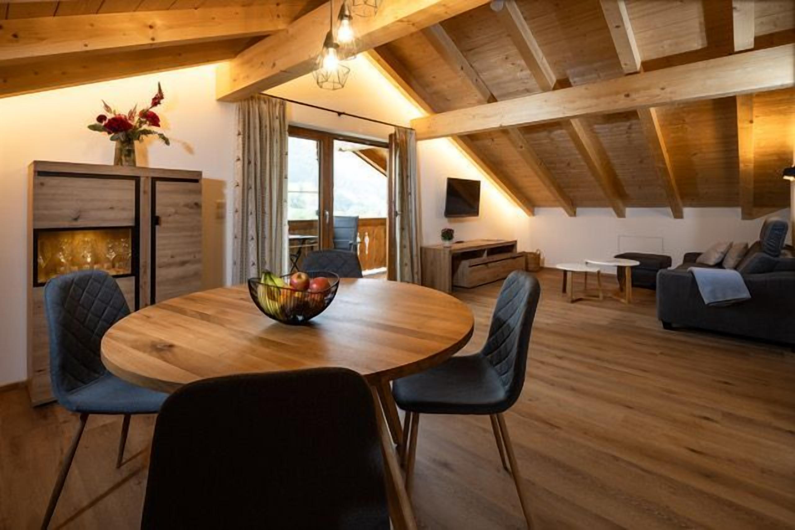 Een eethoek met een ronde tafel, stoelen en een schaal fruit. Achtergrond toont een bank en een televisie.