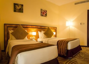 Two beds with headboards and bedside tables, illuminated by wall-mounted lamps, in a room with a carpeted floor and framed artwork.