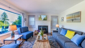 Ein Wohnzimmer mit Kamin, zwei Sofas, einem Couchtisch und Blick auf Bäume durch große Fenster.