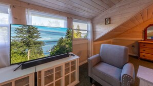 Ein gemütliches Cabin-Zimmer mit einem großen Fernseher auf einer Holzkommode, einem bequemen grauen Sofa und Blick auf einen See und Wald durch die Fenster.
