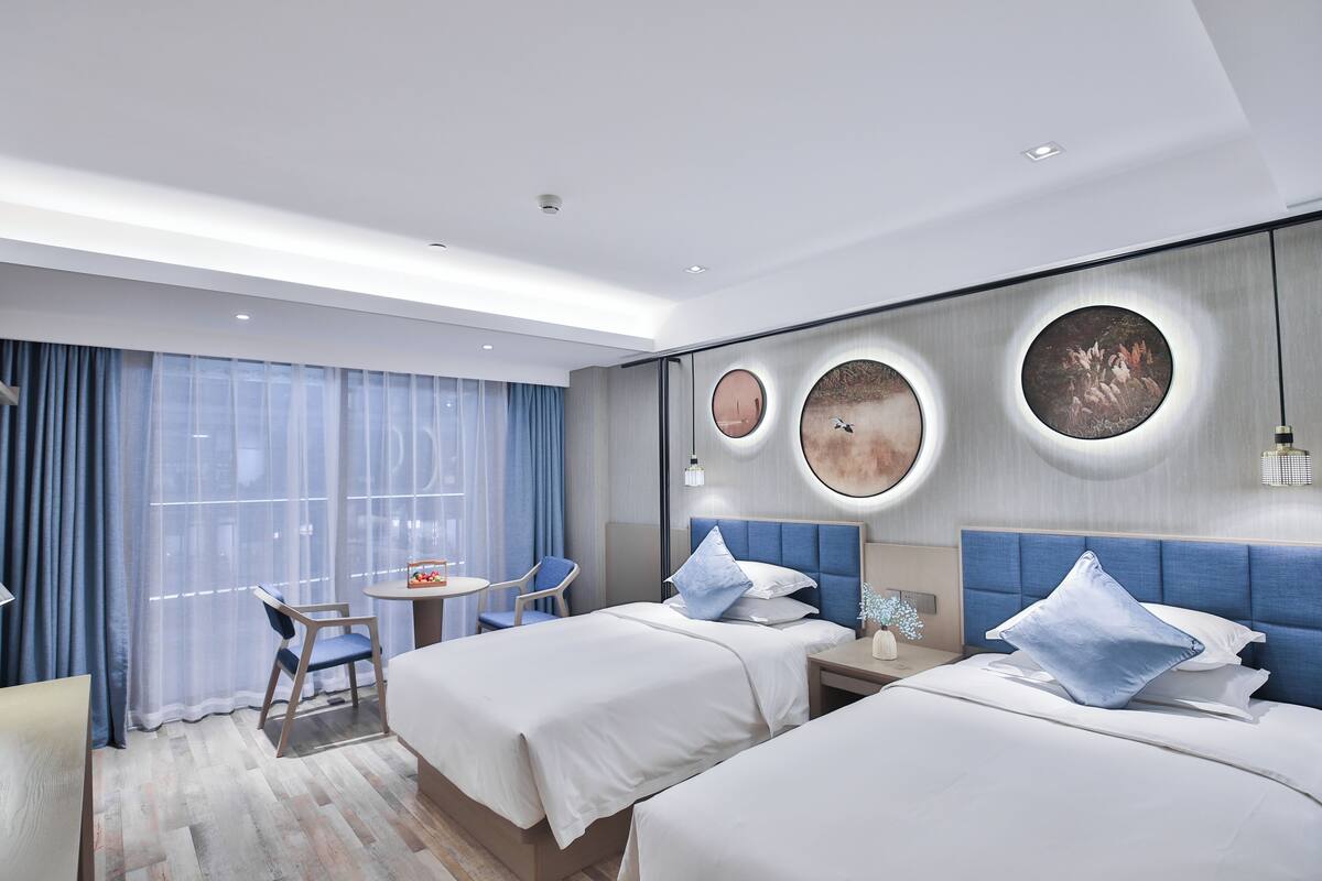 A hotel room with two beds, a small table, a chair, and a large window with curtains.