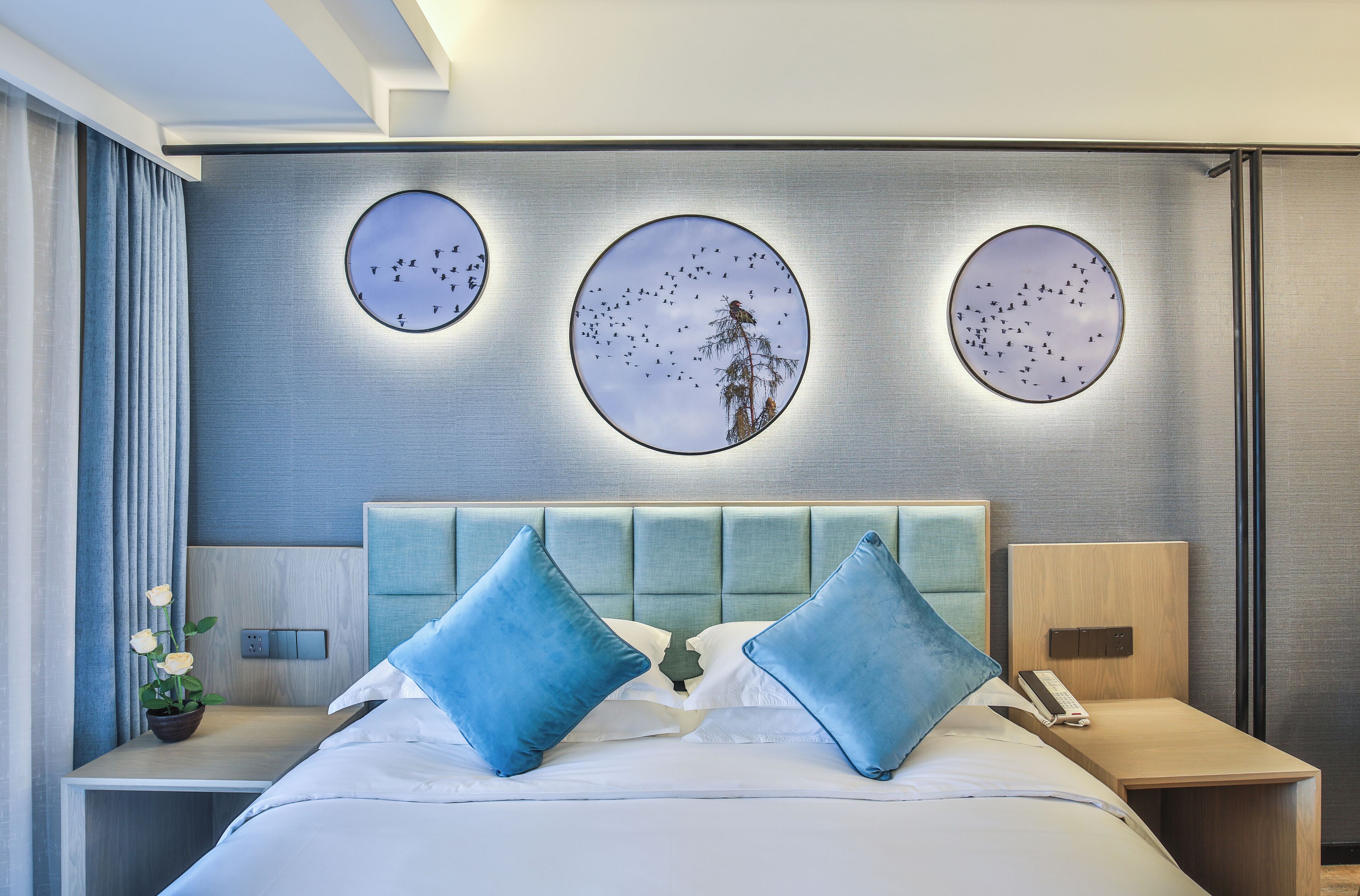 Ein modernes Hotelzimmer mit einem großen Bett, zwei blauen Kissen, einem Nachttisch mit Telefon und drei runden Wandbildern mit Vogelmotiven.