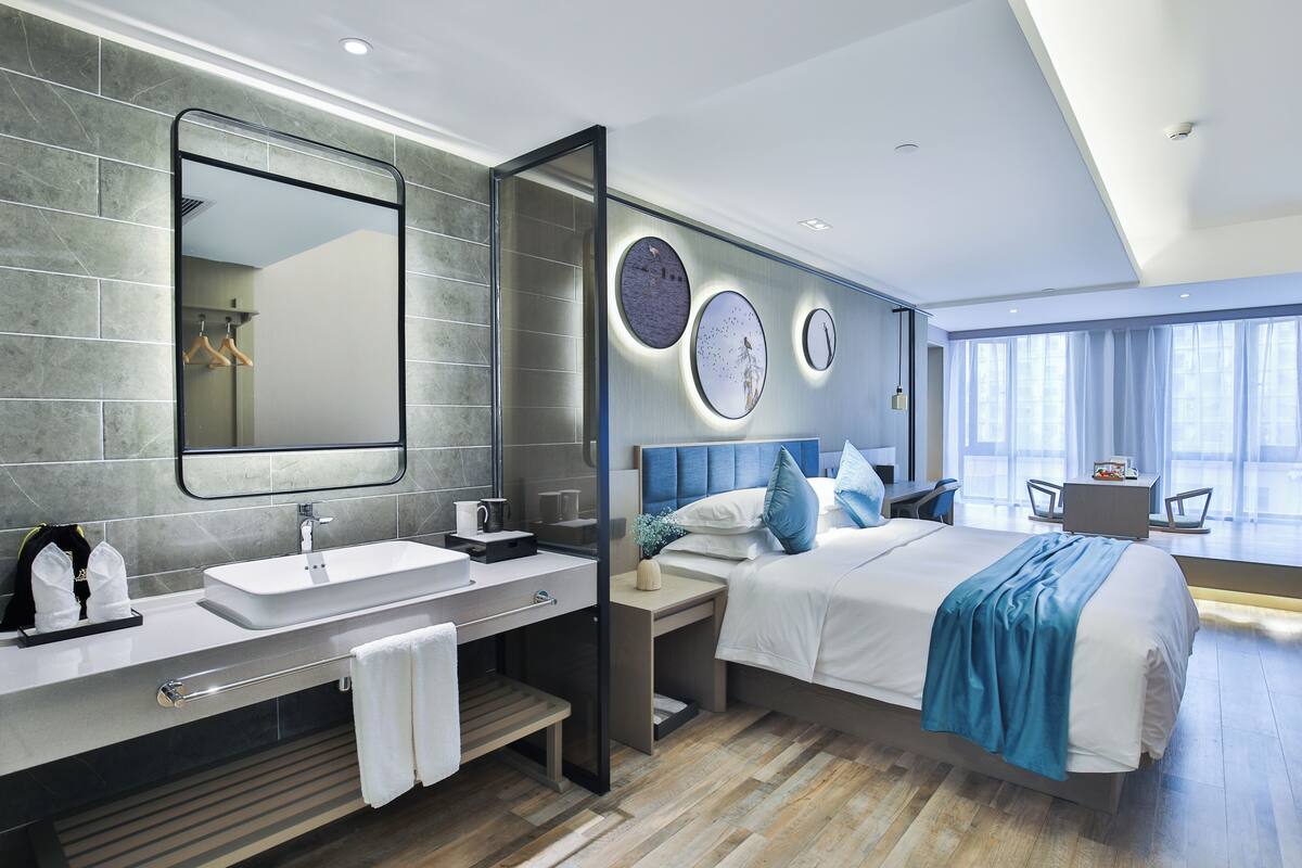 A modern hotel room with a large bed, a bedside table, a vanity with a mirror, and a large window offering a view.