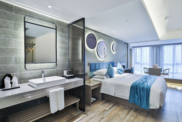 A modern hotel room with a large bed, a bedside table, a vanity with a mirror, and a large window offering a view.